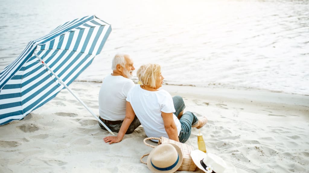 An older couple sits on the sandy Virginia beach under a striped umbrella, facing the water, with hats and a bag placed nearby.